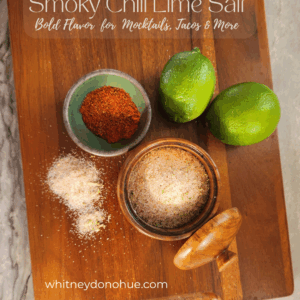 Overhead view of smoky chili lime salt in a wooden salt cellar with limes, chili powder, and spilled seasoning on a wooden board