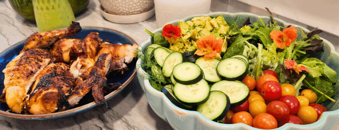 Dinner spread with rotisserie chicken and a colorful spring salad featuring cucumbers, cherry tomatoes, avocado, and edible nasturtiums