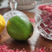A glass jar of raspberry citrus finishing salt on a wooden board, surrounded by scattered salt, a lemon, and a lime
