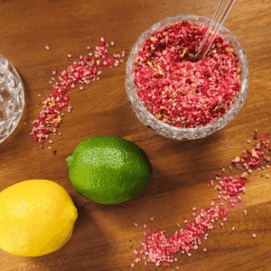 Close-up of raspberry citrus finishing salt in a glass jar with scattered salt, a lemon, and a lime on a wooden surface