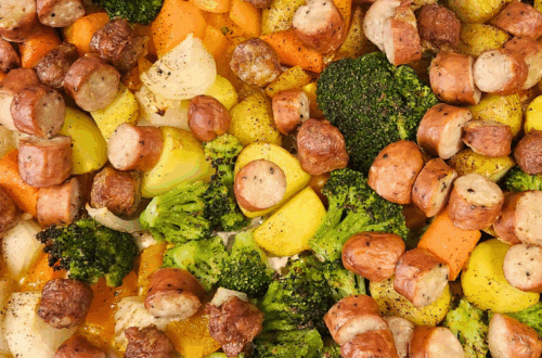 Wide shot of a foil-lined sheet pan piled with roasted sausage slices, baby potatoes, broccoli florets, carrot chunks, and onion pieces