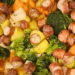Wide shot of a foil-lined sheet pan piled with roasted sausage slices, baby potatoes, broccoli florets, carrot chunks, and onion pieces