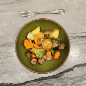 Overhead shot of a green bowl filled with roasted sausage slices, baby potatoes, broccoli florets, and carrot chunks, with a silver fork resting above on a marble countertop