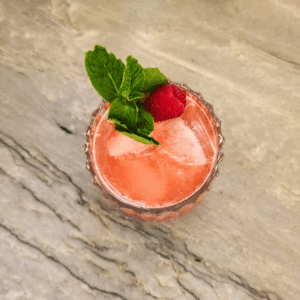 Coral pink Wild May Cup mocktail garnished with mint and a fresh raspberry in a pressed glass