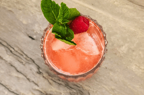 Coral pink Wild May Cup mocktail garnished with mint and a fresh raspberry in a pressed glass