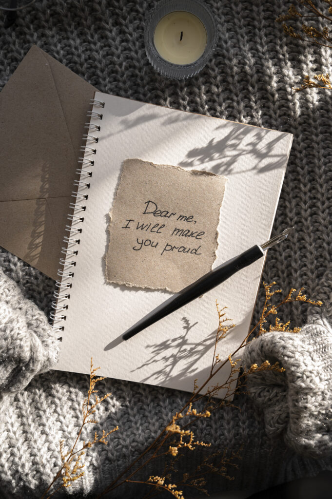 Handwritten note that reads "Dear me, I will make you proud" on torn paper atop a spiral notebook, with a pen, candle, and cozy sweater in soft sunlight.