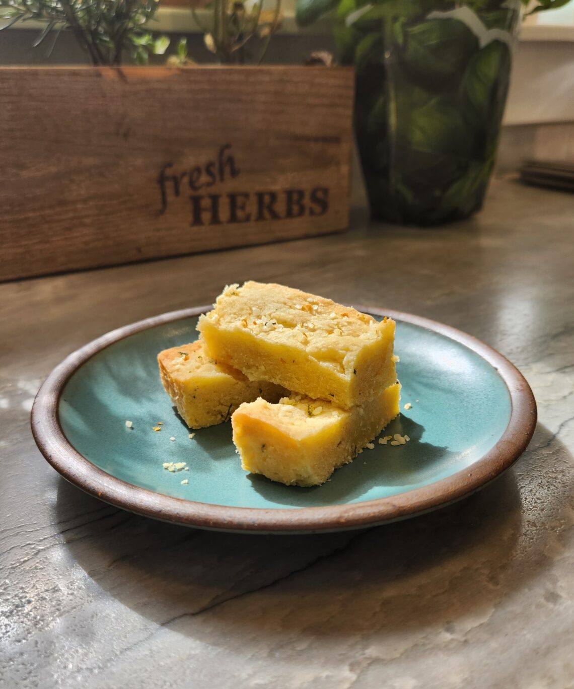 Rosemary lemon shortbread bars stacked on a blue plate with fresh herbs and a “fresh HERBS” wooden box in the background, basking in natural sunlight.