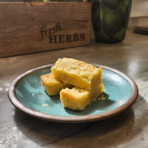 Rosemary lemon shortbread bars stacked on a blue plate with fresh herbs and a “fresh HERBS” wooden box in the background, basking in natural sunlight.