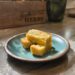 Rosemary lemon shortbread bars stacked on a blue plate with fresh herbs and a “fresh HERBS” wooden box in the background, basking in natural sunlight.