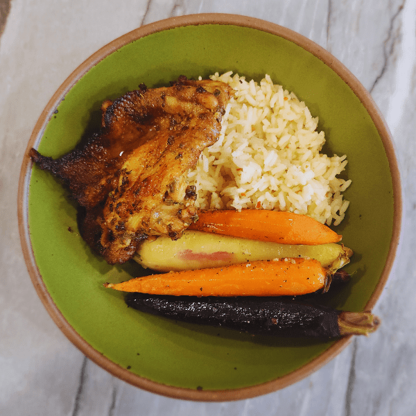 Grilled Cajun chicken thigh served with seasoned rice and colorful roasted carrots in a green bowl.