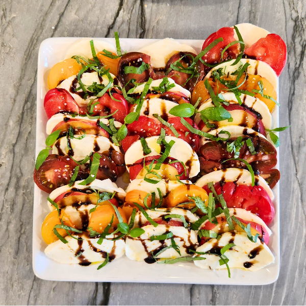 A vibrant caprese salad made with heirloom tomatoes, fresh peaches, mozzarella, basil ribbons, and a drizzle of balsamic glaze, arranged on a white platter.