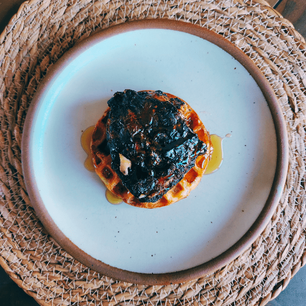 “Grilled chicken thigh on a herbed sourdough waffle, drizzled with honey, served on a ceramic plate over a woven placemat.”