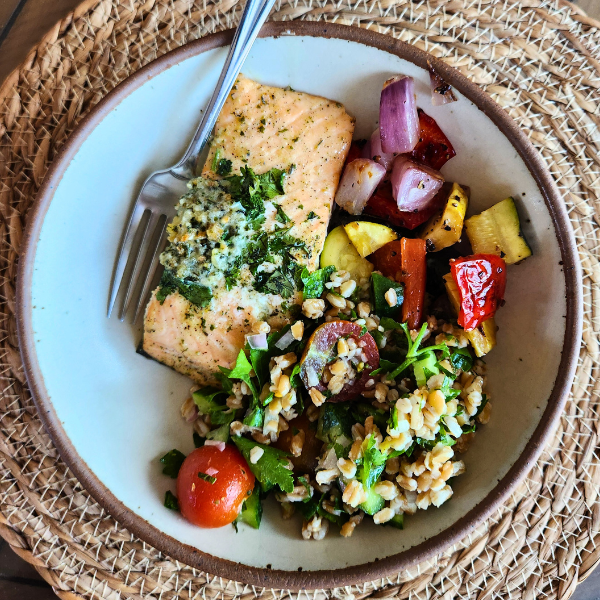 A colorful plate of grilled salmon with herb butter, farro tabbouleh, and roasted summer vegetables, served on a woven charger.