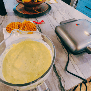 “Overhead view of sourdough waffle batter, a tray of cooked waffles, and a waffle maker on a kitchen counter.”