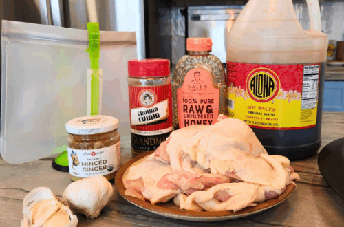 Ingredients for Thai Chicken marinade including soy sauce, honey, ground cumin, fresh garlic and ginger, arranged beside raw chicken thighs on a kitchen counter.
