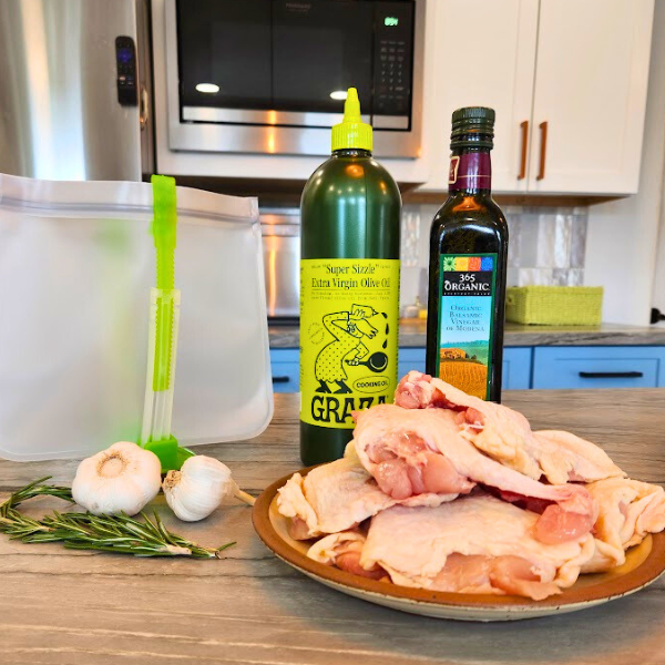 Ingredients for Tuscan Chicken marinade: olive oil, balsamic vinegar, rosemary, garlic, and raw chicken thighs arranged on a kitchen counter with a reusable freezer bag.