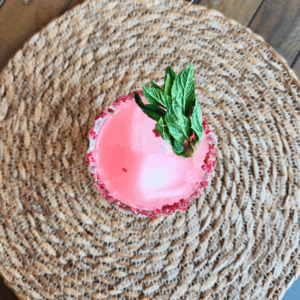Overhead view of a pink Witchlight Fizz mocktail in a faceted glass, rimmed with raspberry citrus salt and garnished with a fresh mint sprig, placed on a woven placemat.