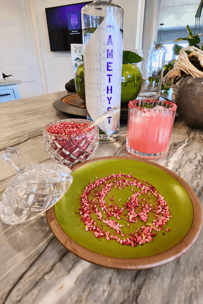 A crystal jar of raspberry citrus salt, a green plate with salt for rimming, a bottle of Amethyst Blueberry Mint, and a prepared Witchlight Fizz mocktail on a marble countertop.