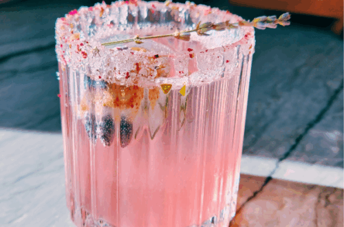 Pink floral mocktail in a crystal glass with a sugared lavender and rose petal rim, garnished with a lavender sprig, glowing in natural sunlight on a marble countertop.