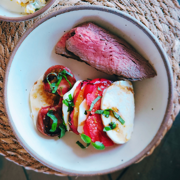 Sliced Santa Maria-style tri-tip on a cutting board, grilled medium-rare with visible sear marks. Fresh heirloom tomatoes and peaches are arranged beside the beef.
