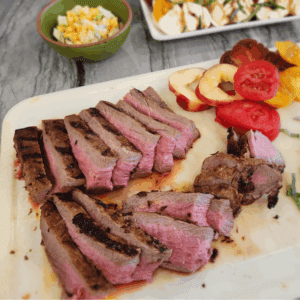 Sliced Santa Maria-style tri-tip on a cutting board, grilled medium-rare with visible sear marks. Fresh heirloom tomatoes and peaches are arranged beside the beef.